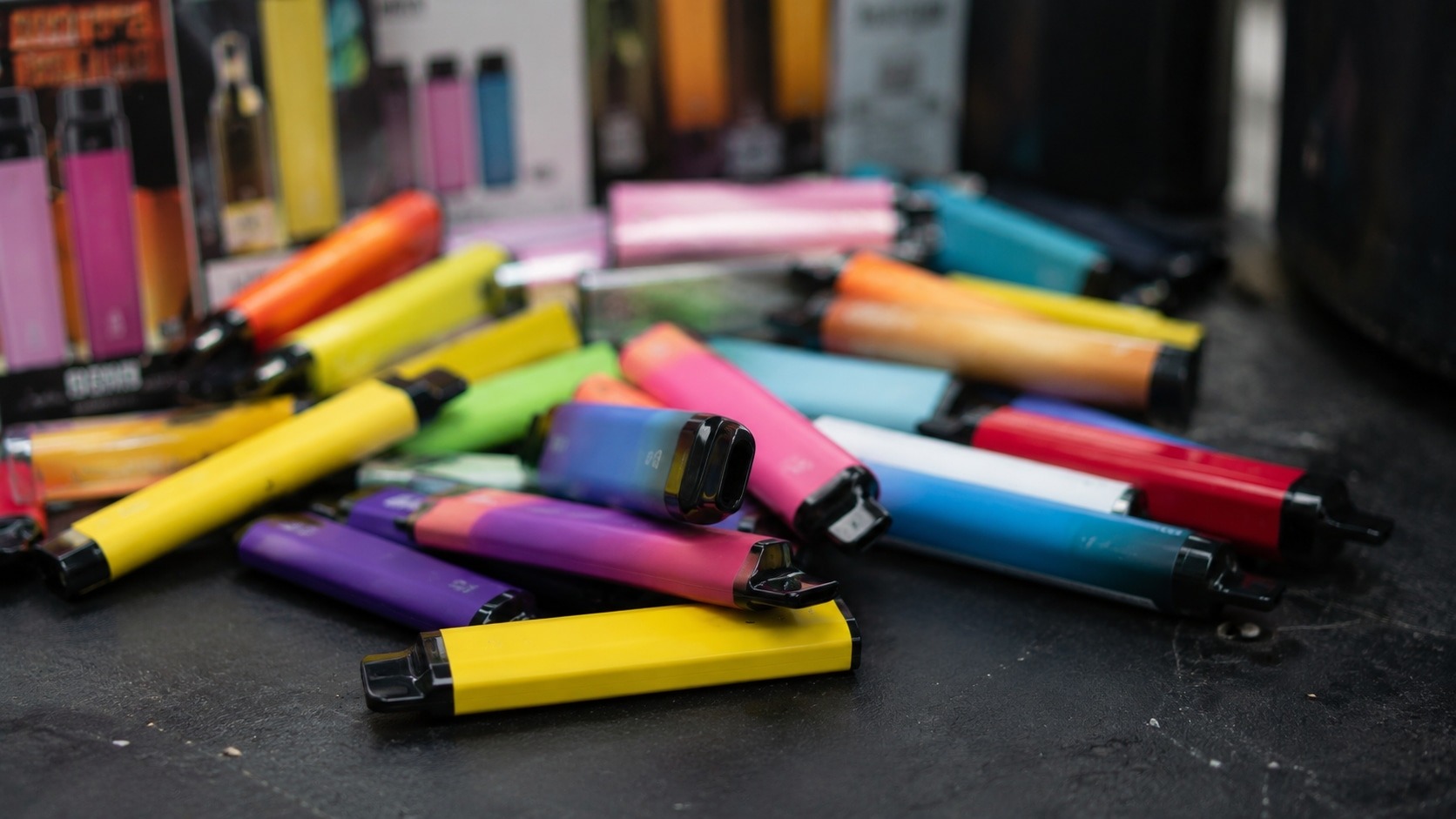 Des dizaines de cigarettes électroniques jetables de couleurs vives amassées sur un sol sombre.